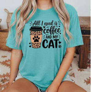 May include: A turquoise t-shirt with the slogan "All I need is coffee and my cat." The design features a coffee cup with a leopard print and paw prints. The shirt has short sleeves.