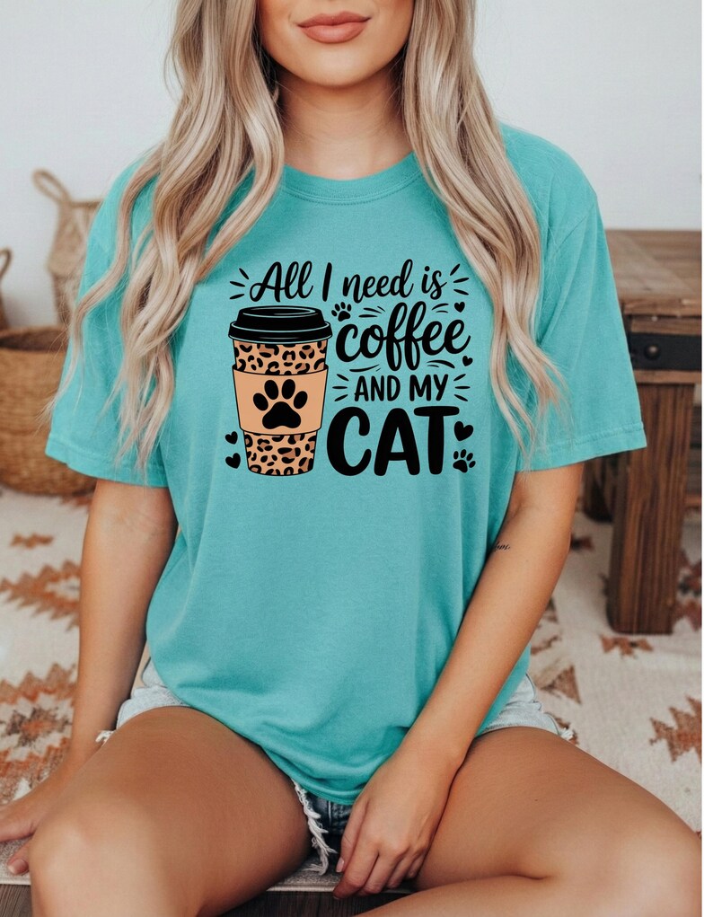 May include: A turquoise t-shirt with the slogan "All I need is coffee and my cat." The design features a coffee cup with a leopard print and paw prints. The shirt has short sleeves.