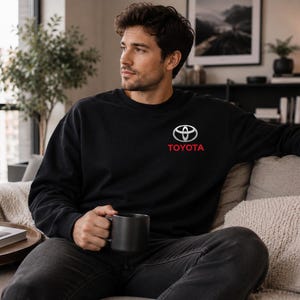 May include: Black sweatshirt with the Toyota logo in white and red. The sweatshirt is long-sleeved and has a crew neck. The person in the image is holding a black mug.