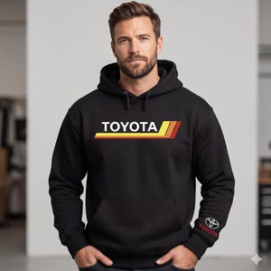 May include: A black hooded sweatshirt featuring the white Toyota logo centered on the chest above horizontal yellow, orange, and red stripes. The left sleeve cuff displays a small Toyota emblem and the word Toyota in red lettering.