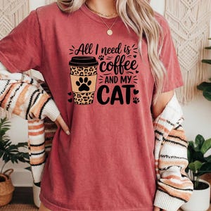May include: A maroon t-shirt with the slogan "All I need is coffee and my cat." The design features a coffee cup with a leopard print and a paw print. The shirt is short-sleeved.