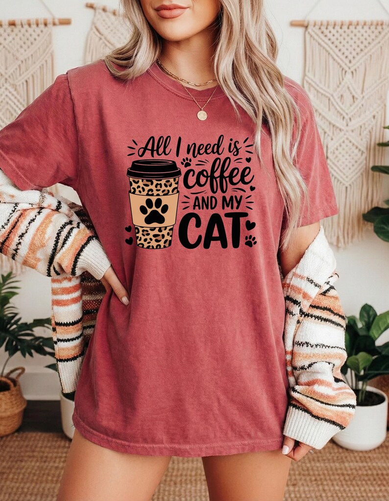 May include: A maroon t-shirt with the slogan "All I need is coffee and my cat." The design features a coffee cup with a leopard print and a paw print. The shirt is short-sleeved.