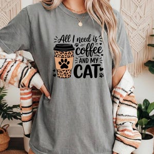 May include: Grey t-shirt featuring the text "All I need is coffee and my cat." The design includes a coffee cup with a leopard print and a cat paw print. The shirt is short-sleeved.