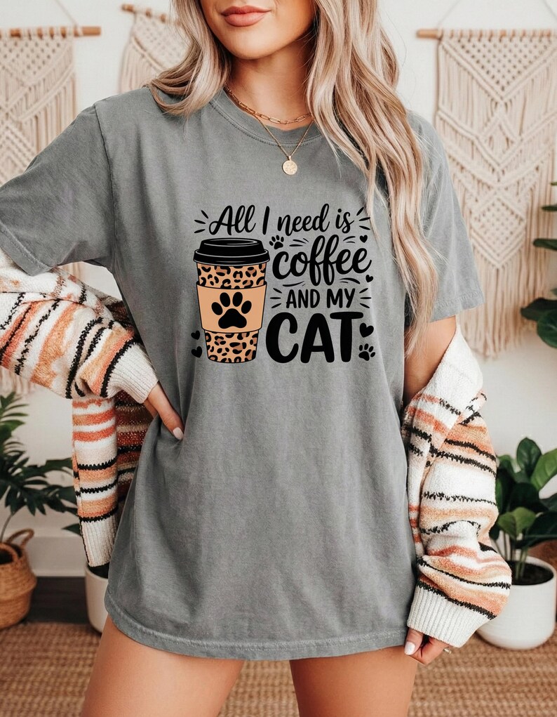 May include: Grey t-shirt featuring the text "All I need is coffee and my cat." The design includes a coffee cup with a leopard print and a cat paw print. The shirt is short-sleeved.