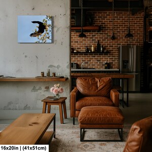 May include: A framed canvas print showing a black eagle in flight against a pale blue sky, on a flowering branch. The artwork measures 41x51cm. The print is displayed in a modern living space with leather furniture and a brick wall.