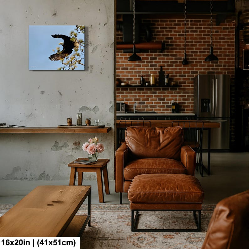 May include: A framed canvas print showing a black eagle in flight against a pale blue sky, on a flowering branch. The artwork measures 41x51cm. The print is displayed in a modern living space with leather furniture and a brick wall.