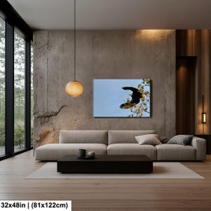 May include: A canvas print of a flying eagle against a blue sky, displayed above a sofa in a modern living room. The artwork measures 81x122cm. The room has a minimalist design with a concrete wall and wooden flooring.