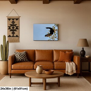 May include: A framed canvas print showing a black bird in flight against a blue sky, with white flowers. The artwork is displayed above a brown leather sofa. The print measures 61x91cm.