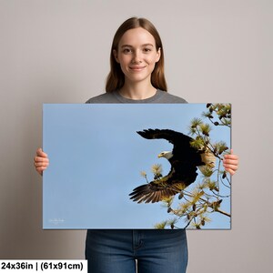 May include: A canvas print showing a bald eagle in flight against a blue sky. The eagle, with its white head and dark wings, is perched on a pine branch. The print measures 61x91cm.