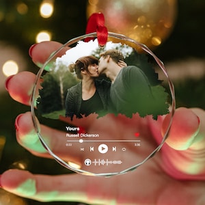 May include: A clear, round ornament with a photo of a kissing couple. The ornament has a red ribbon for hanging and features a music player interface with the song title "Yours" by Russell Dickerson. The ornament is held in a hand.