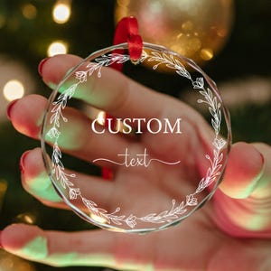 May include: A clear, round ornament with a white floral design around the edge. The words "CUSTOM text" are printed in the center. A red ribbon is attached for hanging. The ornament is held in front of a blurred background.