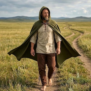 May include: A person wearing a green hooded cloak with a leather clasp, a beige shirt, and brown pants. The person is walking on a dirt path through a grassy field. The cloak is flowing in the wind.