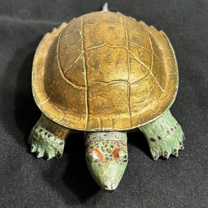 May include: A vintage turtle figurine with a golden-brown shell and green head and feet. The shell has a textured, segmented pattern. The turtle's head features red and white accents. The figurine is likely made of metal.