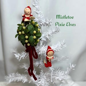 May include: A white Christmas tree adorned with two vintage-style pixie elf ornaments and a mistletoe decoration. The elves wear red outfits and hats. The mistletoe has green leaves, yellow berries, and a red velvet ribbon. Text reads "Mistletoe Pixie Elves."