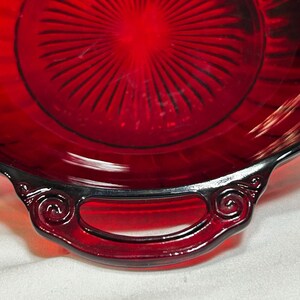 May include: A close-up of a ruby red glass serving dish with a handle. The dish has a circular design with radiating lines and a decorative handle with a swirl design. The glass is glossy and reflects light.