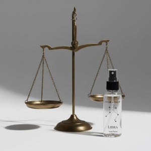 May include: A vintage brass scale stands beside a clear glass spray bottle. The bottle is labeled "STAR + SALT" and "LIBRA Aura Spray 2oz" with a constellation graphic. The scale has two round pans suspended by chains.