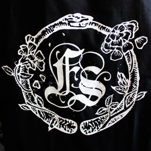 以下が含まれることがあります： 白黒のグラフィックデザイン。スタイリッシュな花輪の中に「FS」の文字が書かれている。