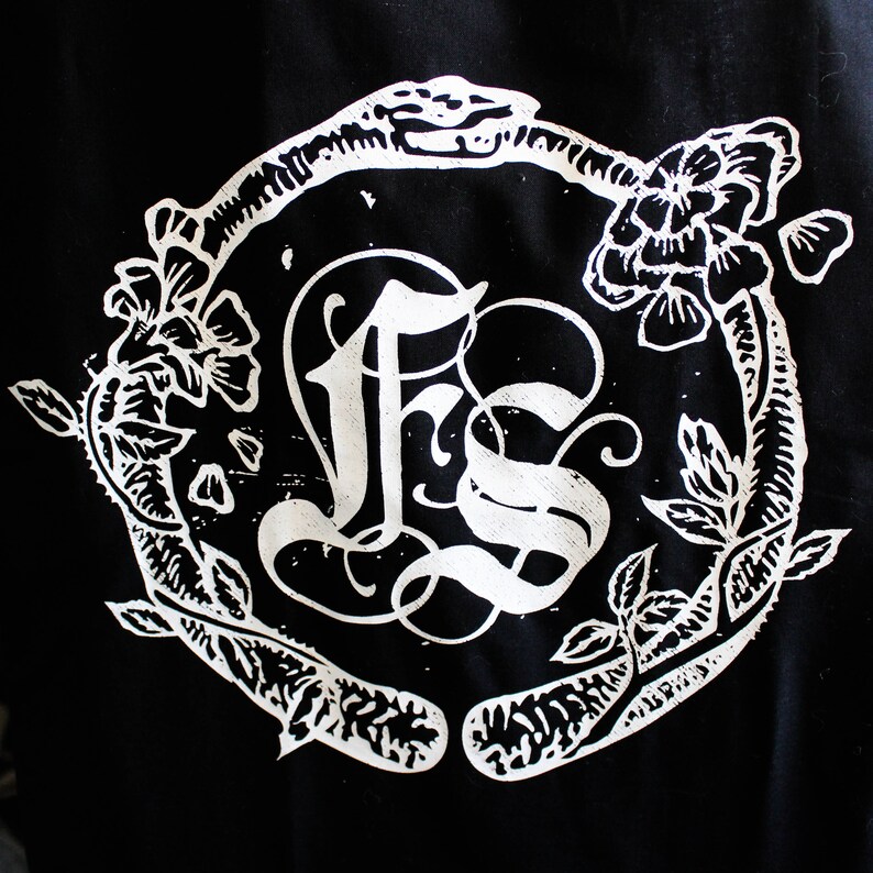 以下が含まれることがあります： 白黒のグラフィックデザイン。スタイリッシュな花輪の中に「FS」の文字が書かれている。