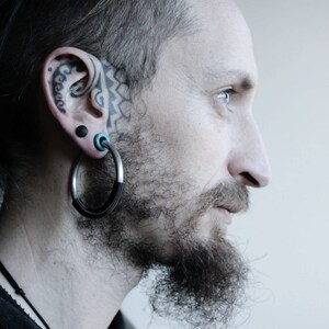May include: A man with a beard and a tattoo on his ear, wearing large hoop earrings. The earrings are made of silver and have a black cord wrapped around them.