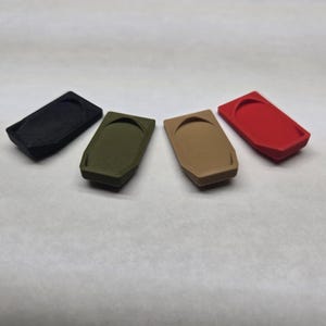 Pode incluir: Quatro objetos retangulares texturizados em preto, verde-oliva, bege e vermelho, dispostos sobre uma superfície branca. Cada um tem uma reentrância curva. Podem ser pequenos recipientes.