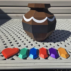 May include: A brown, geometric vase with white curved accents and a black band. Below are a red heart and six colorful, faceted gems in green, blue, red, purple, and yellow. The objects are arranged on a white, patterned surface.