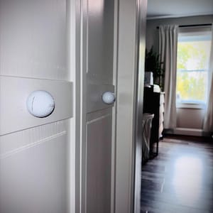 May include: A light grey door with a golf ball used as a door knob. The golf ball is attached to a rectangular panel on the door. The door is slightly ajar, revealing a glimpse of a room. The door is painted a light grey colour.