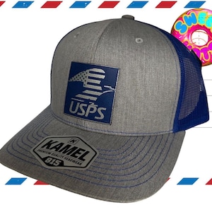 May include: A gray and blue trucker hat featuring a square patch with the USPS eagle logo on the front. The hat has a gray heathered front panel, a blue mesh back, and a sticker on the brim that reads KAMEL PREMIUM QUALITY HEADWEAR 815.
