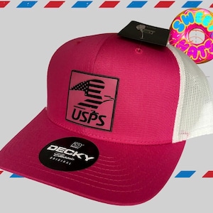 Op de afbeelding: Een felroze snapback-pet met een wit mesh achterpaneel. De voorkant is voorzien van een zwarte vierkante patch met een gestileerde adelaar en de tekst USPS. Een zwarte ronde sticker op de platte klep bevat de tekst DECKY Classic ORIGINAL.
