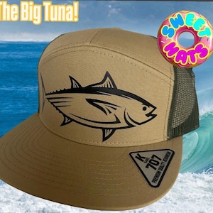 May include: A tan trucker hat featuring a black tuna fish design. The hat has a mesh back and a flat peak. The text "The Big Tuna!" is above the hat, and a donut with the text "Sweet Hats" is to the right.