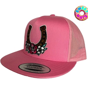 May include: Pink trucker hat with a horseshoe and floral embroidery. The hat features a curved brim, a mesh back, and an adjustable snapback closure. The horseshoe is brown with red and white flowers.