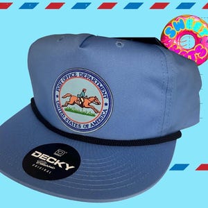 Könnte beinhalten: Eine hellblaue Baseballkappe mit einer schwarzen Kordelverzierung an der Basis der Krone. Die Vorderseite zeigt einen runden Aufnäher mit einem Reiter auf einem Pferd und dem Text Post Office Department United States of America.