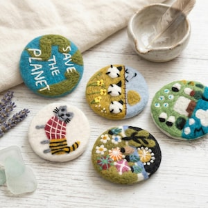 Pode incluir: Cinco emblemas circulares de lã feltrada com vários desenhos: uma terra azul e verde com o texto SAVE THE PLANET, um gato com uma camisa xadrez vermelha, uma paisagem com ovelhas, um jardim floral e um padrão abstrato azul e verde.