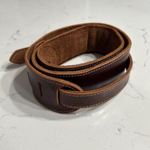 May include: A rolled-up brown leather guitar strap with white stitching. The strap has a soft, suede-like interior and a buckle for adjustment. The leather is a rich, dark brown color.