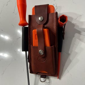 May include: A brown leather tool belt with a metal clasp, holding several orange-handled tools, including a screwdriver and a wrench. The belt also has a black holder for a metal tool and a D-ring at the bottom.