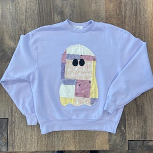 May include: A light purple sweatshirt with a patchwork ghost design on the front. The ghost has black oval eyes and is made of various fabric patterns in pastel colors. The sweatshirt has long sleeves and a crew neck.