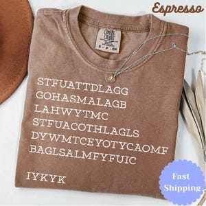 Puede incluir: Camiseta color espresso con texto blanco. La camiseta tiene cuello redondo y mangas cortas. Un collar plateado con un peque&ntilde;o colgante est&aacute; drapeado sobre la camiseta. El texto de la camiseta incluye el acr&oacute;nimo "IYKYK".