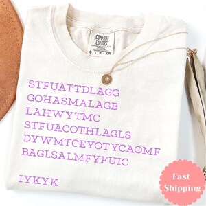 Puede incluir: Camiseta Comfort Colors color crema con texto morado y collar dorado. El texto incluye el acr&oacute;nimo "IYKYK" y otras palabras. Tambi&eacute;n es visible una pegatina rosa con las palabras "Fast Shipping".