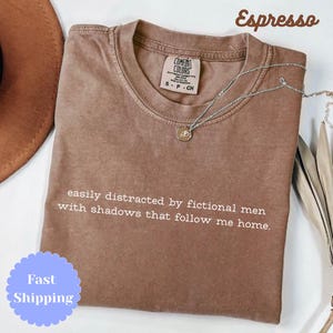 May include: A brown t-shirt with the text "easily distracted by fictional men with shadows that follow me home." The shirt is labelled "Espresso" and has a silver necklace with a pendant. The shirt has a Comfort Colors tag.