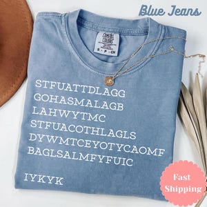 Puede incluir: Una camiseta Comfort Colors azul claro con texto blanco y un collar dorado. La camiseta tiene el texto "STFUATTDLAGG GOHASMALAGB LAHWYTMC STFUACOTHLAGLS DYWMTCEYOTYCAOMF BAGLSALMFYFUIC IYKYK" impreso. Tambi&eacute;n se ven las palabras "Blue Jeans".