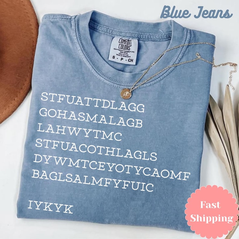 Puede incluir: Una camiseta Comfort Colors azul claro con texto blanco y un collar dorado. La camiseta tiene el texto "STFUATTDLAGG GOHASMALAGB LAHWYTMC STFUACOTHLAGLS DYWMTCEYOTYCAOMF BAGLSALMFYFUIC IYKYK" impreso. Tambi&eacute;n se ven las palabras "Blue Jeans".
