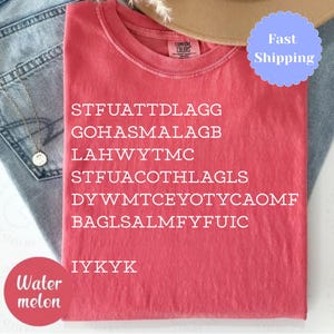 Puede incluir: Una camiseta roja con texto blanco, incluyendo el acr&oacute;nimo "IYKYK". La camiseta est&aacute; doblada y colocada sobre unos vaqueros azules. Tambi&eacute;n se ven un collar dorado y un sombrero beige.