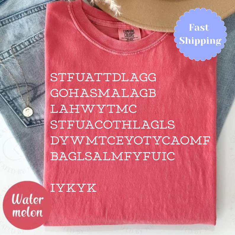 Puede incluir: Una camiseta roja con texto blanco, incluyendo el acr&oacute;nimo "IYKYK". La camiseta est&aacute; doblada y colocada sobre unos vaqueros azules. Tambi&eacute;n se ven un collar dorado y un sombrero beige.