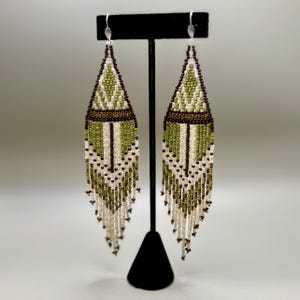 Könnte beinhalten: Ein Paar Perlenohrringe mit geometrischem Design. Die Ohrringe zeigen eine Kombination aus olivgrünen, braunen und cremefarbenen Perlen. Sie werden auf einem schwarzen Ständer vor einem neutralen Hintergrund präsentiert. Die Ohrringe haben Fransen am unteren Ende.