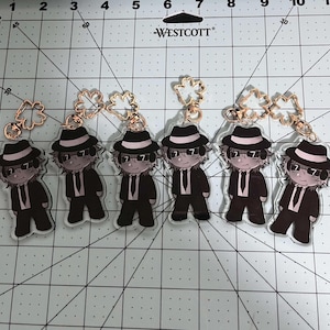 May include: Six acrylic keychains featuring a cartoon character in a black suit, hat, and sunglasses. Each keychain has a gold-colored clasp. The background is a grid.