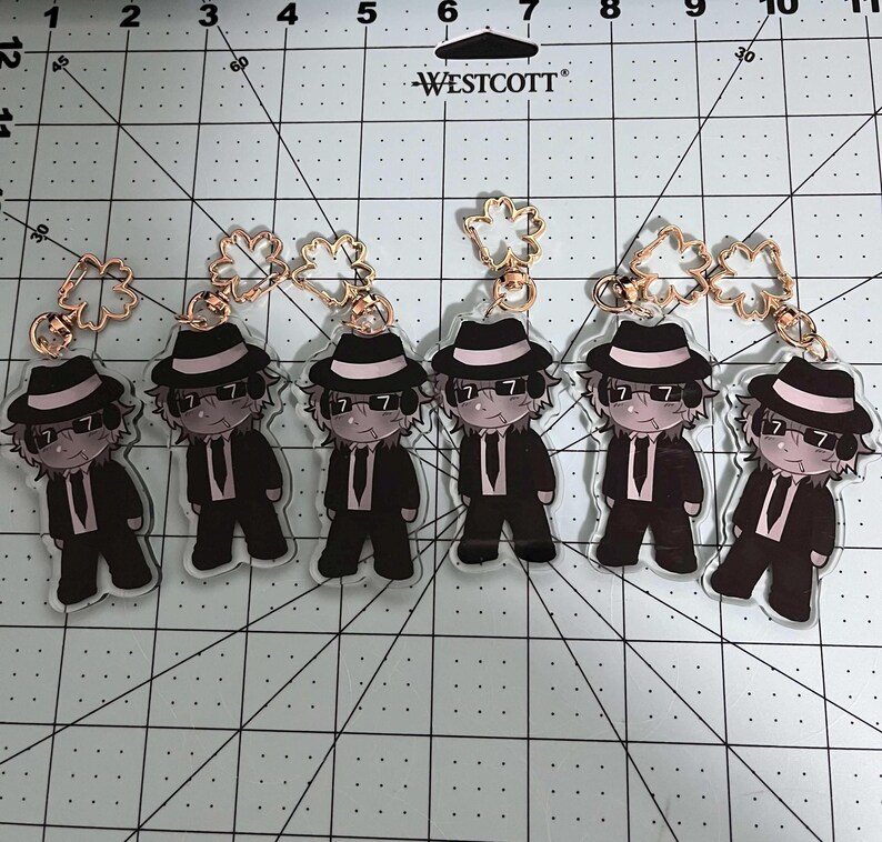 May include: Six acrylic keychains featuring a cartoon character in a black suit, hat, and sunglasses. Each keychain has a gold-colored clasp. The background is a grid.
