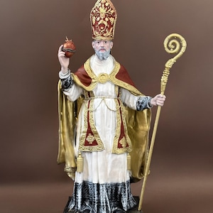 Op de afbeelding: Gedetailleerd beeld van een religieuze figuur, met een rode en gouden mijter en cape over een wit gewaad. De figuur houdt een staf en een klein, hartvormig voorwerp vast. Het beeld heeft een baard en staat op een donkere basis.