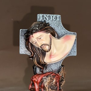 May include: A religious figurine of Jesus on a cross. The gray cross has the letters INRI at the top. Jesus has long brown hair and a beard, with a crown of thorns. The base is a brown rock formation with red paint.