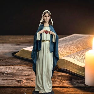 May include: A religious figurine of the Virgin Mary, dressed in a white gown and blue cloak, stands before an open book and a lit candle. The statue has a red heart on her chest. The scene is set on a wooden surface.