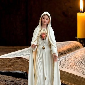 Op de afbeelding: Een wit beeld van een religieuze figuur met een hartvormige hanger, gekleed in een witte jurk en sluier. Een brandende kaars en een open boek staan op de achtergrond. Het beeld staat op een houten oppervlak.