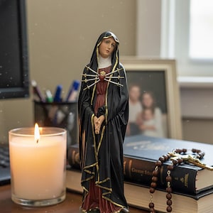 Könnte beinhalten: Eine Statue der Jungfrau Maria, bekleidet mit einem schwarzen Gewand mit goldenen Verzierungen, steht auf einem Schreibtisch. Ein Herz, durchbohrt von sieben Schwertern, befindet sich auf ihrer Brust. Eine brennende Kerze, Bücher und ein Rosenkranz sind ebenfalls auf dem Schreibtisch.